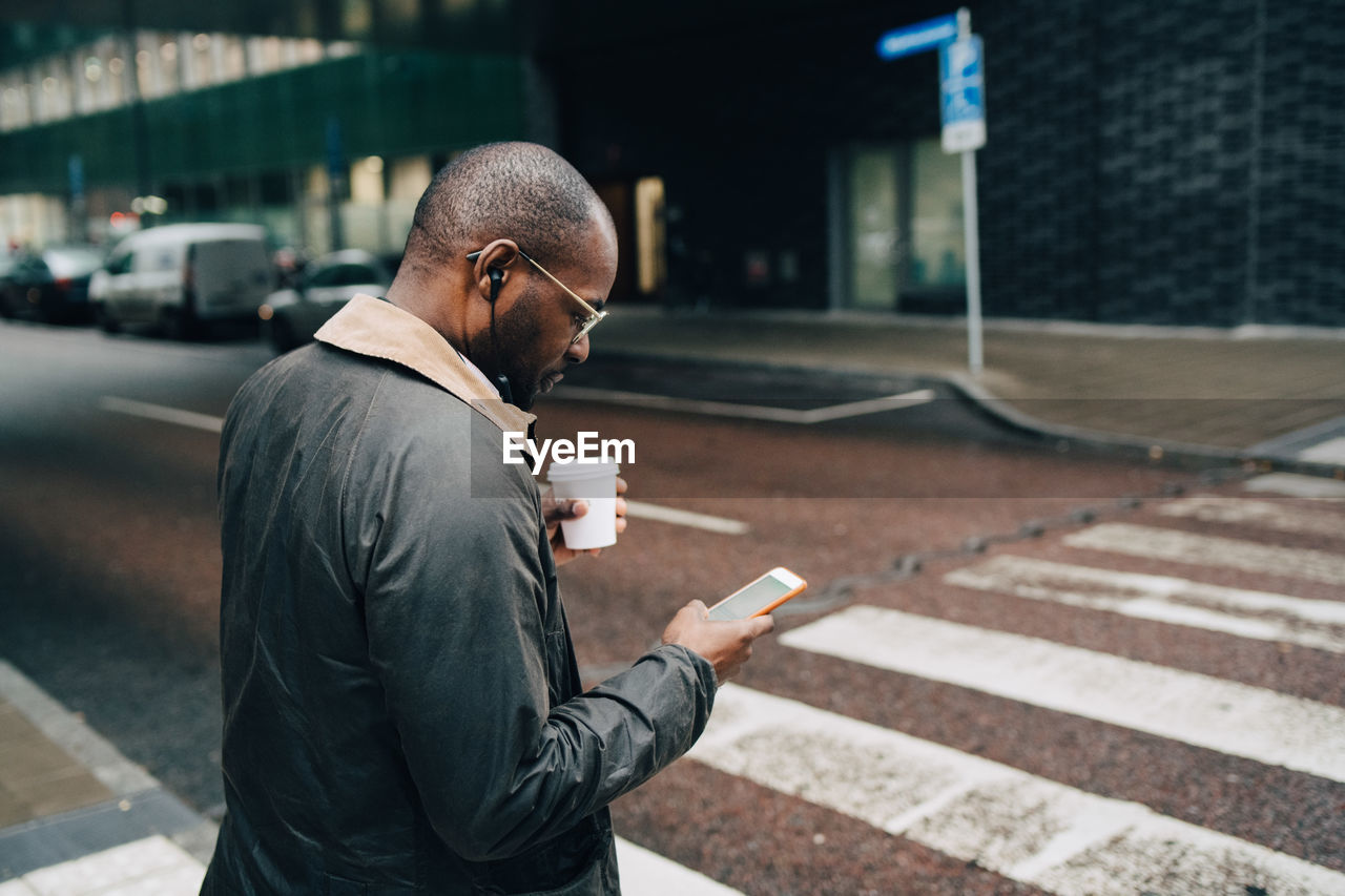 Side view of businessman with coffee using mobile phone while crossing road in city