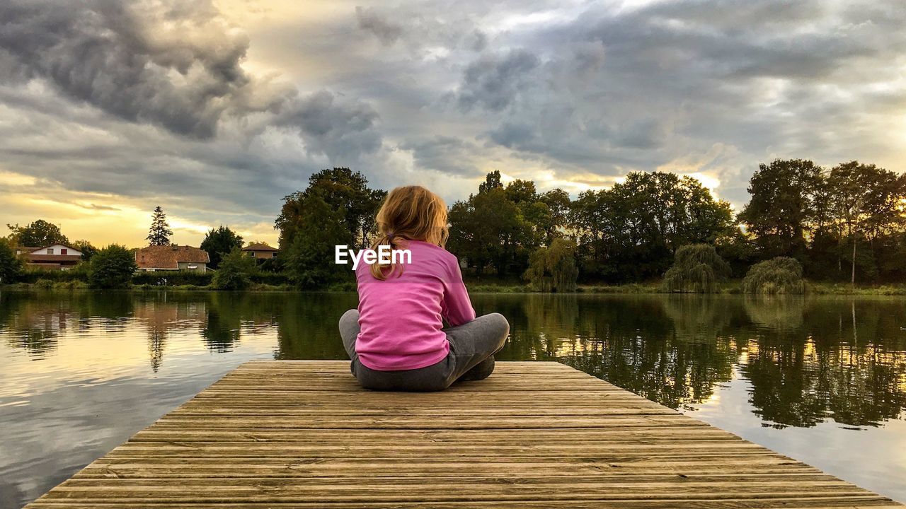 Rear view of girl sitting on pier over lake against sky