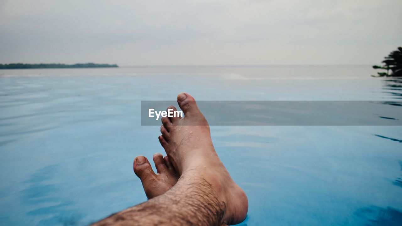 Low section of man relaxing in infinity pool against sea during sunset