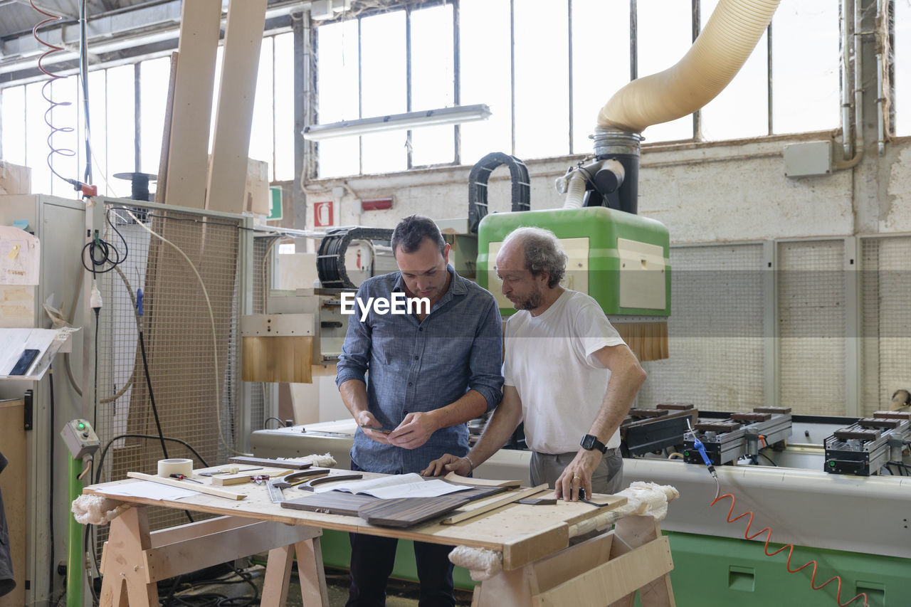 Project manager and coworker working while standing against sawing machine at factory