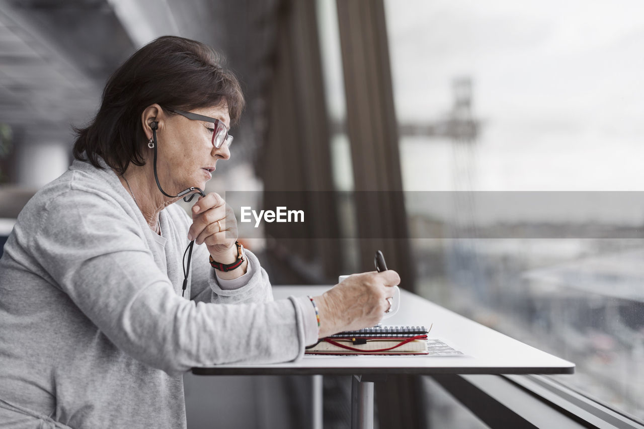 Side view of senior businesswoman using hands-free device by window in office