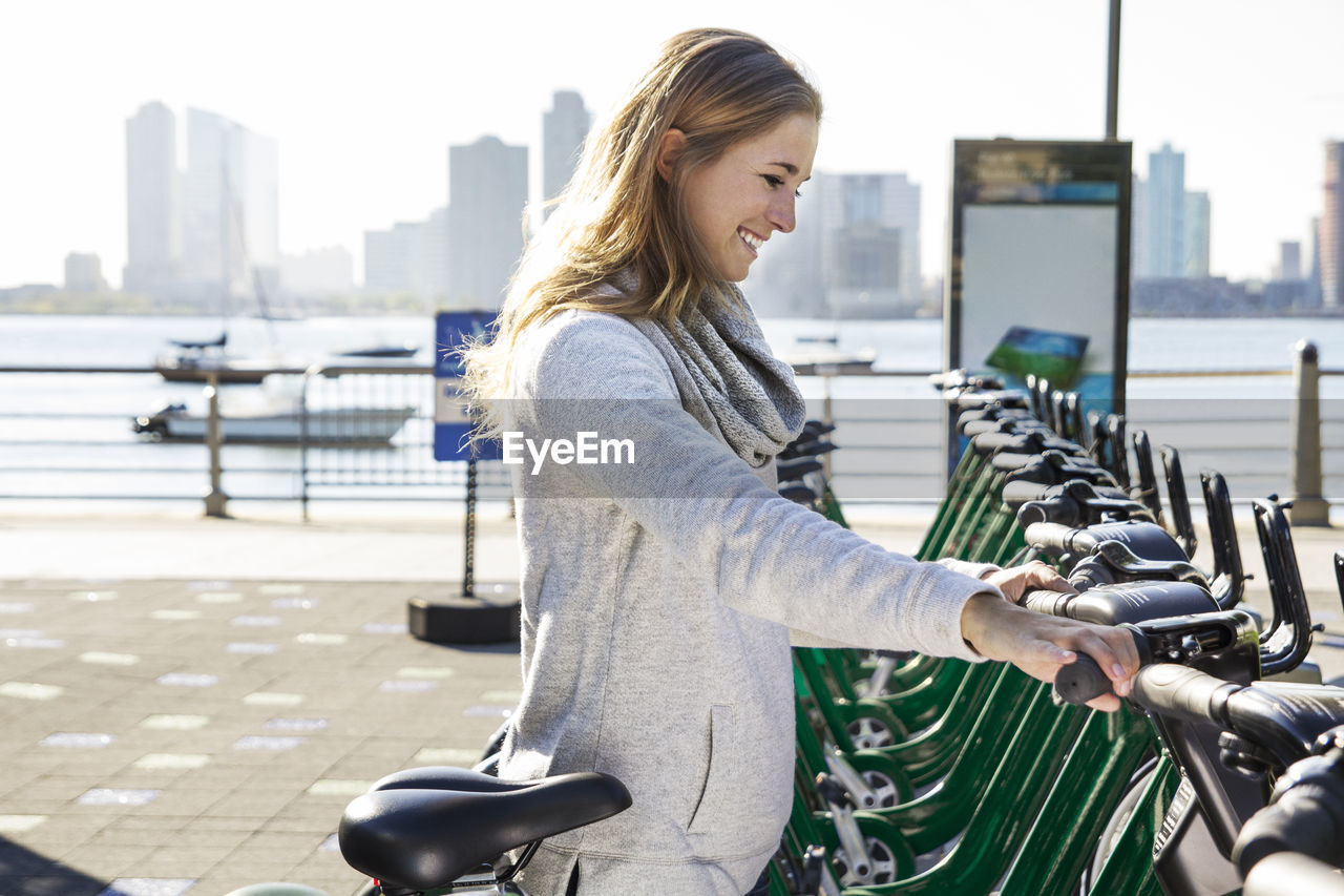 Side view of smiling young woman renting bicycle by lake in city