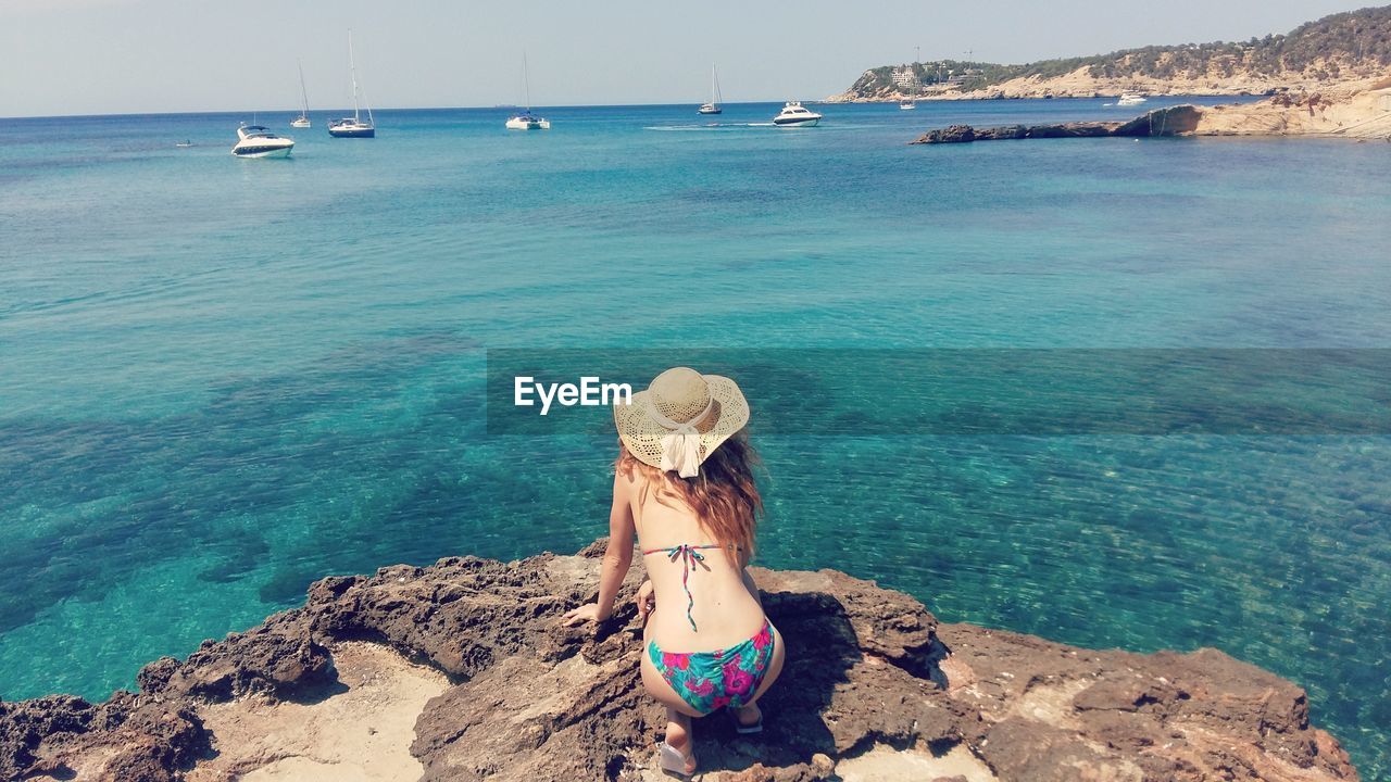 Rear view of woman wearing bikini while kneeling on rocks at beach