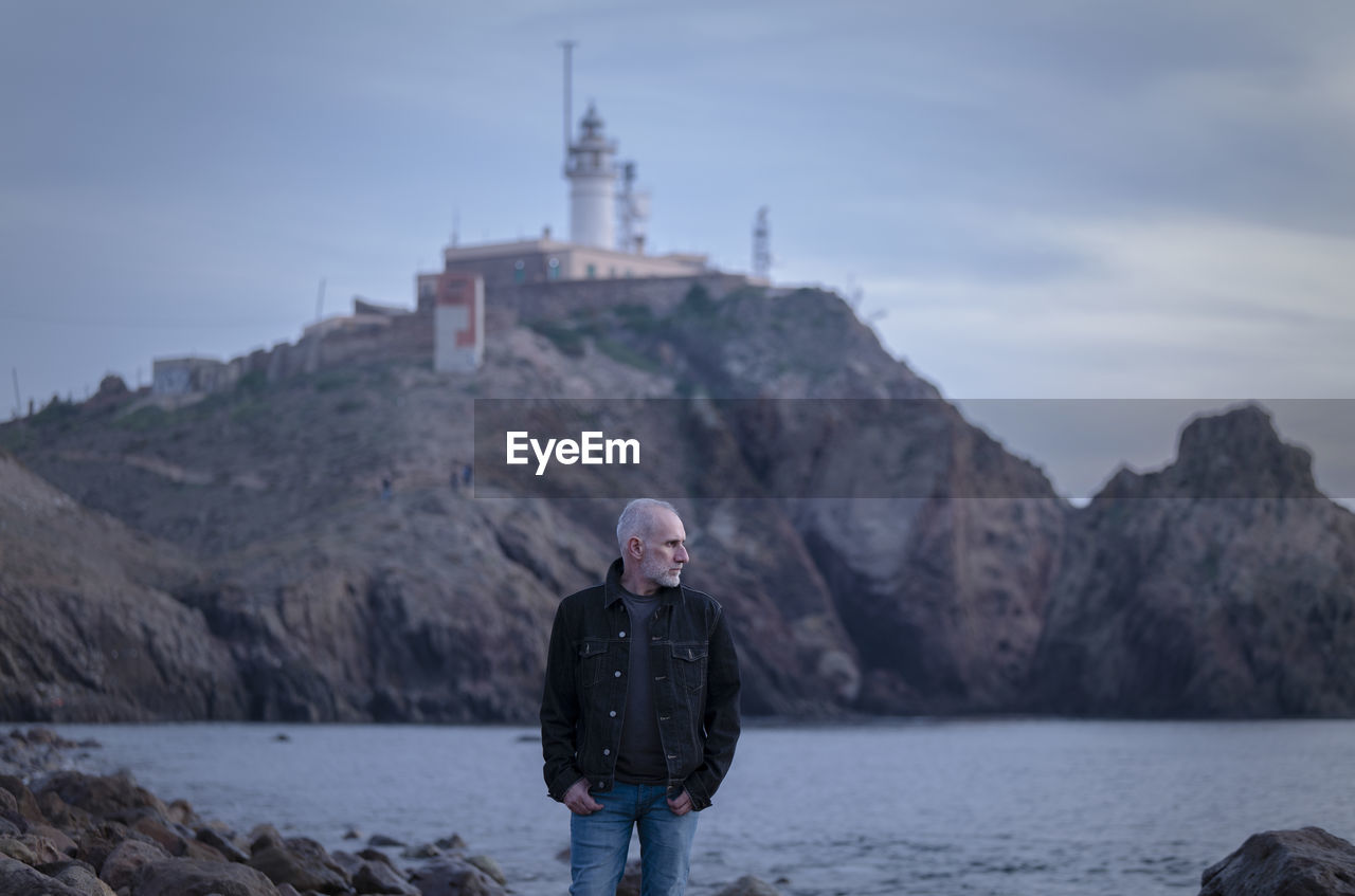 Adult man in black jacket standing on rocky beach looking at sea with lighthouse. almeria, spain
