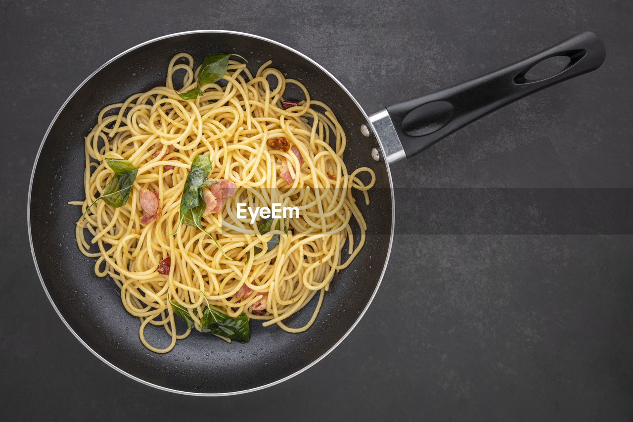 Spaghetti pasta with dried chilli, garlic, sweet basil and bacon in black pan, top view