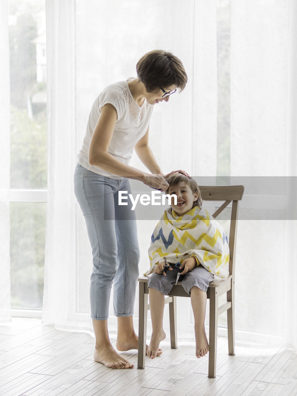 Mother cuts her son's hair by herself. boy sits, covered with cloth, and holds pair of scissors. 