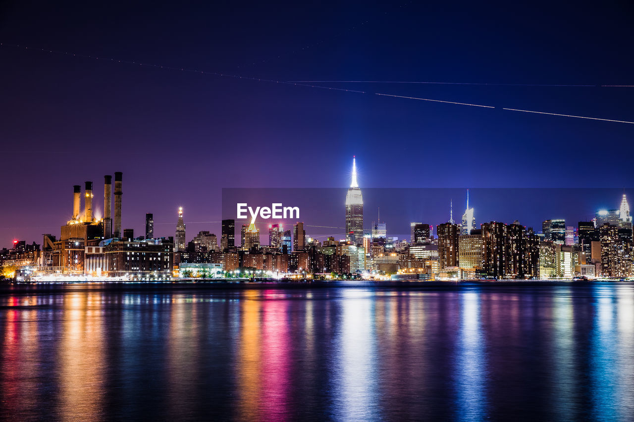 Illuminated buildings in city by river against clear sky at night