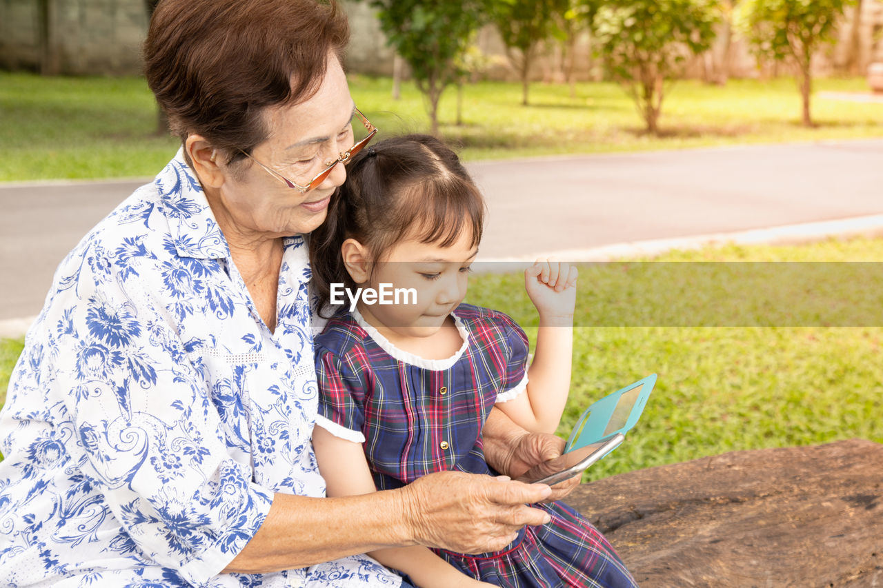 Grandmother using smart phone while sitting with granddaughter at park