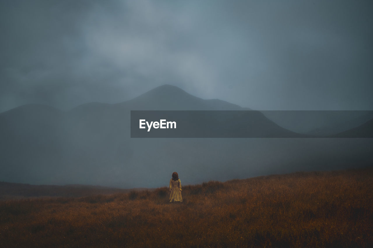 Rear view of woman standing on field against mountain during foggy weather