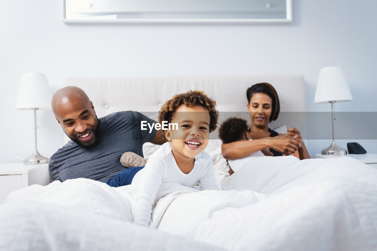 Parents playing with children while relaxing on bed against wall at home