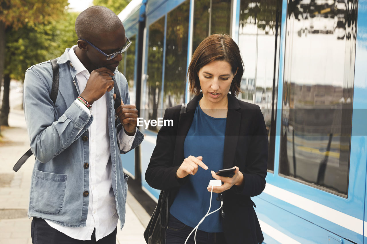 Multi-ethnic commuters discussing over smart phone while standing blue tram in city