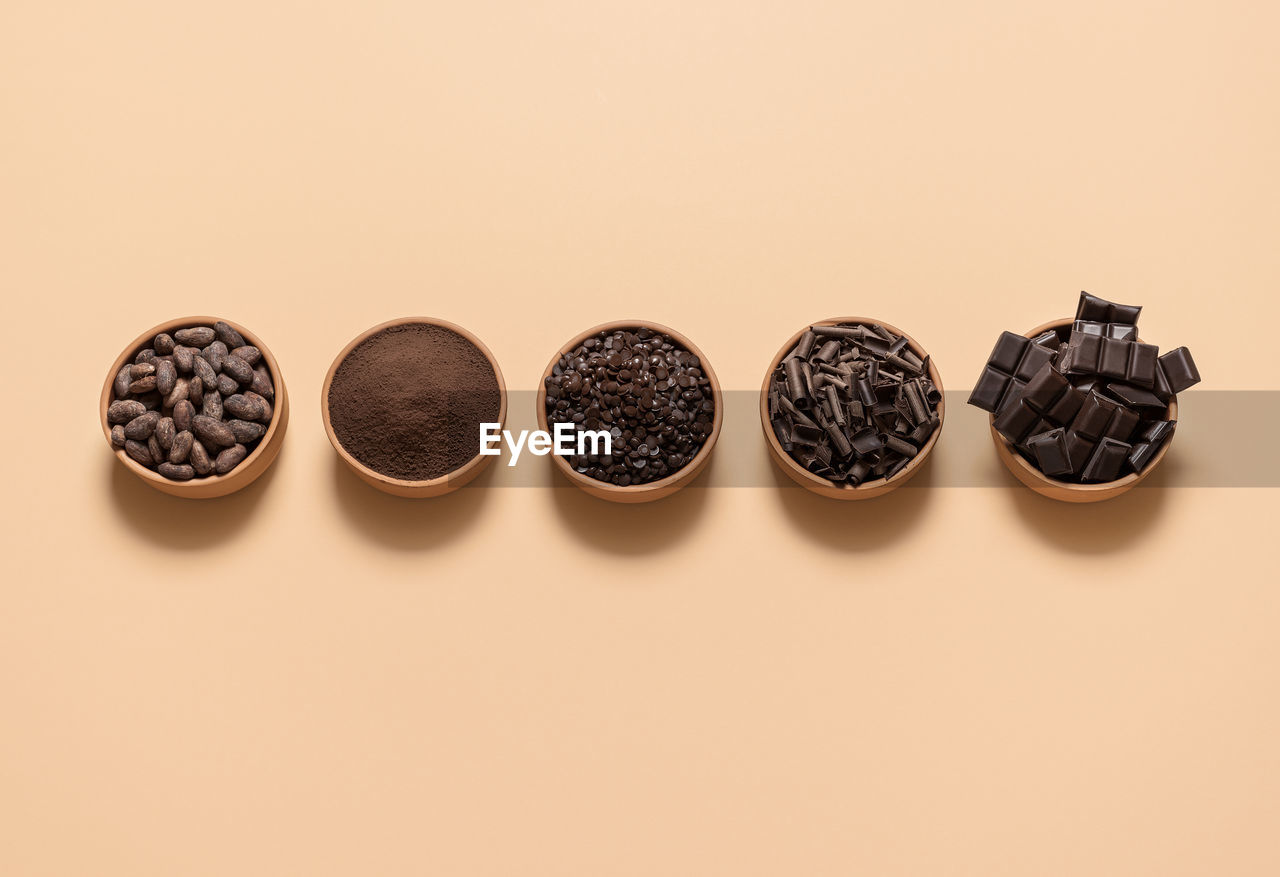Chocolate ingredients, cacao beans, cocoa powder, chocolate chunks in bowls, on a beige background.