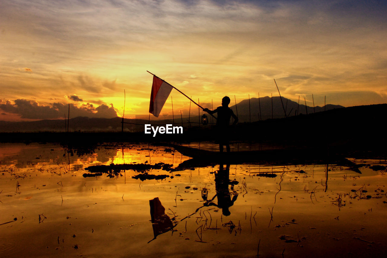 Silhouette man holding flag in lake against sky during sunset