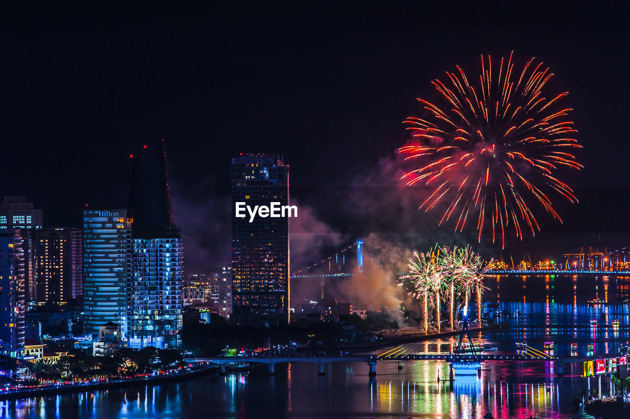 Firework display over illuminated buildings and river in city at night