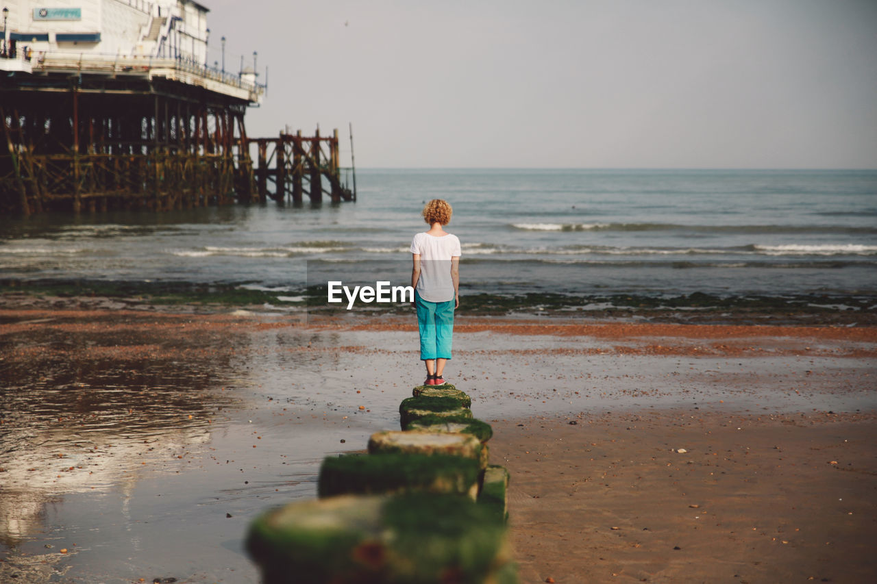 Rear view of woman standing on rock by eastbourne pier at beach against sky