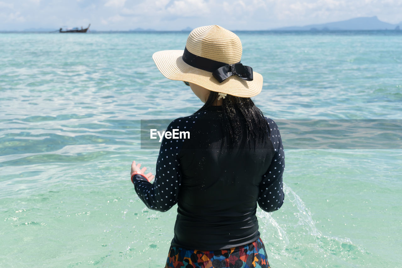 REAR VIEW OF WOMAN WEARING HAT STANDING BY SEA