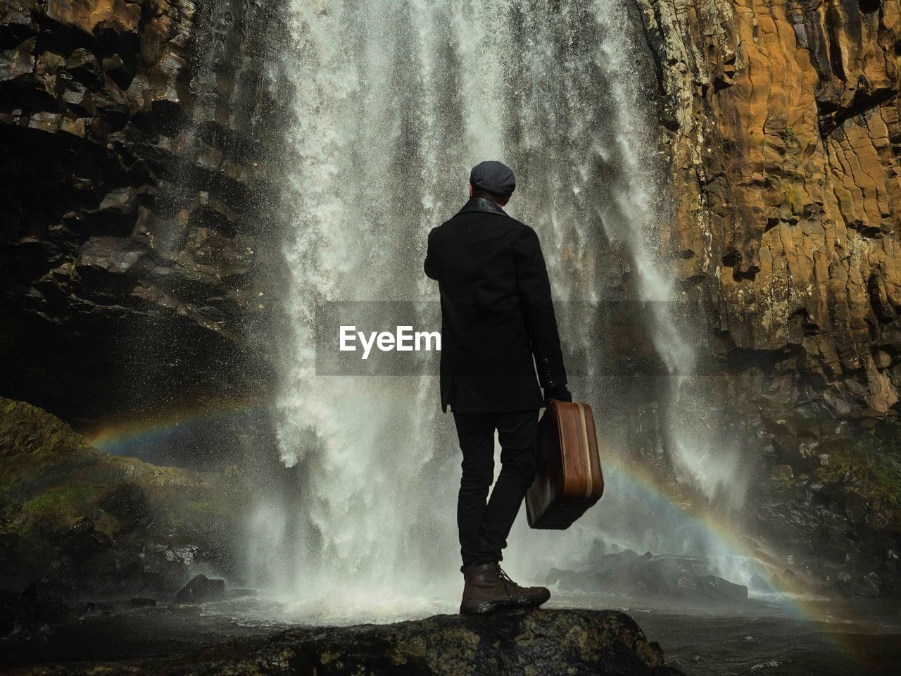 Rear view of man standing on rock while holding briefcase against waterfall