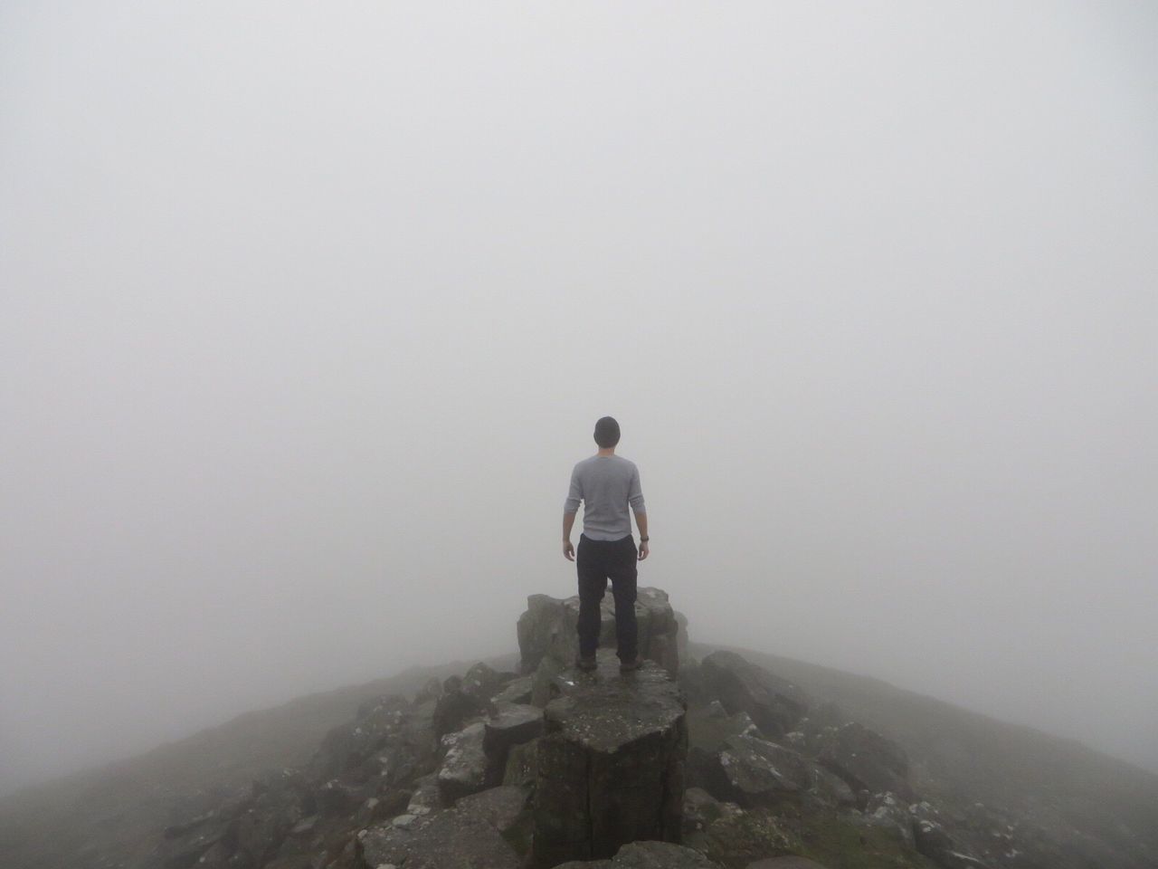 REAR VIEW OF MAN STANDING ON ROCK IN FOGGY WEATHER AGAINST SKY