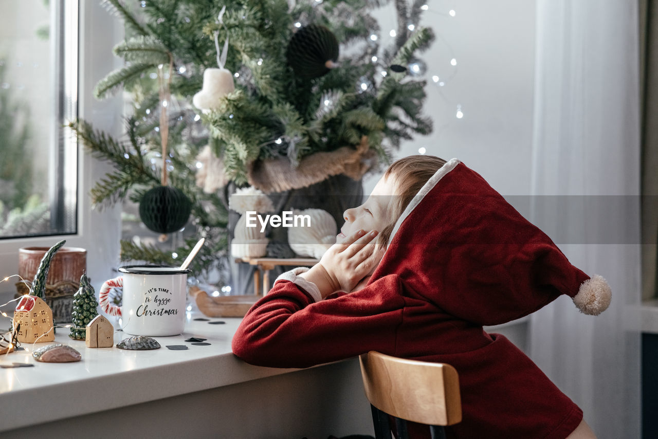 A thoughtful boy in a santa costume sits at the window and makes christmas decorations