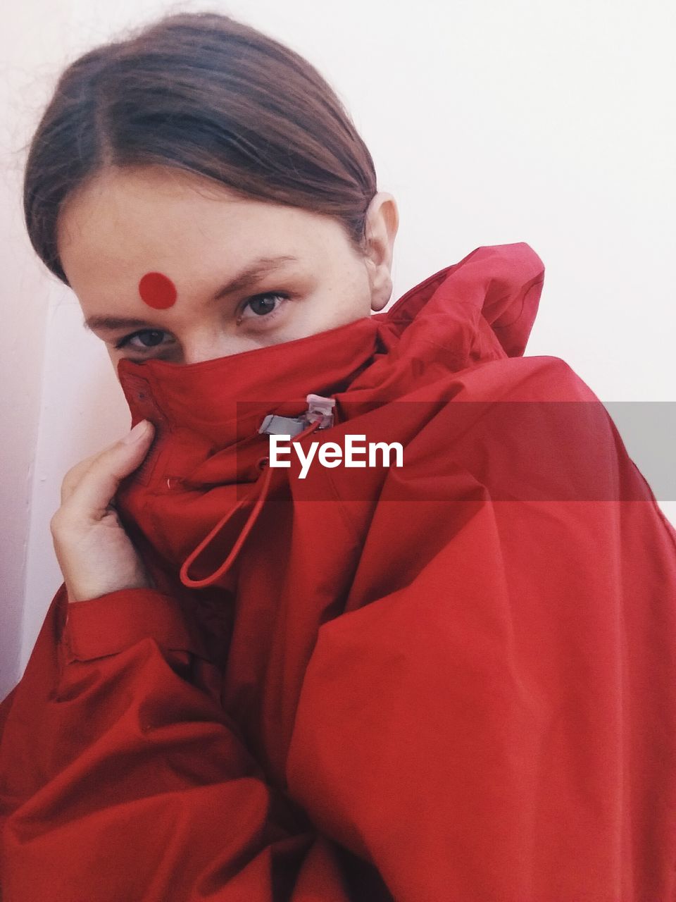 Portrait of woman covering her face in red jacket against white wall