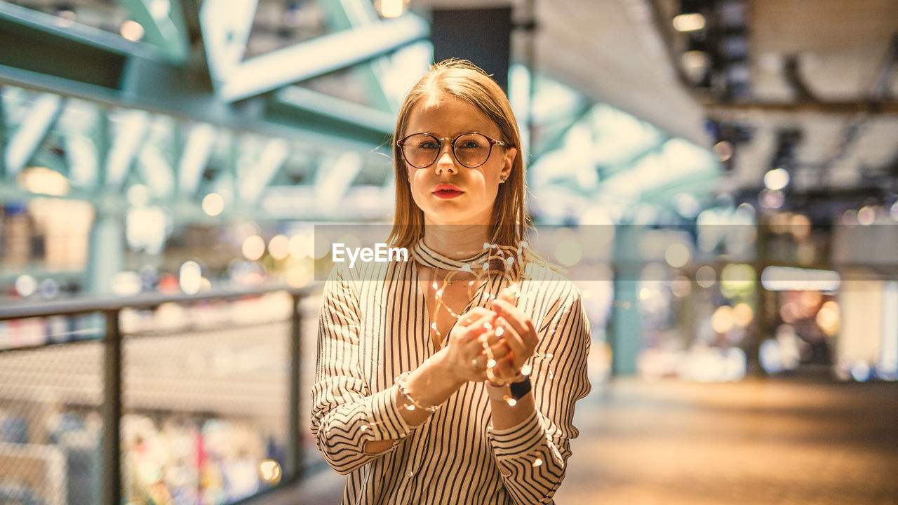 Portrait of young woman holding illuminated string lights while standing in mall