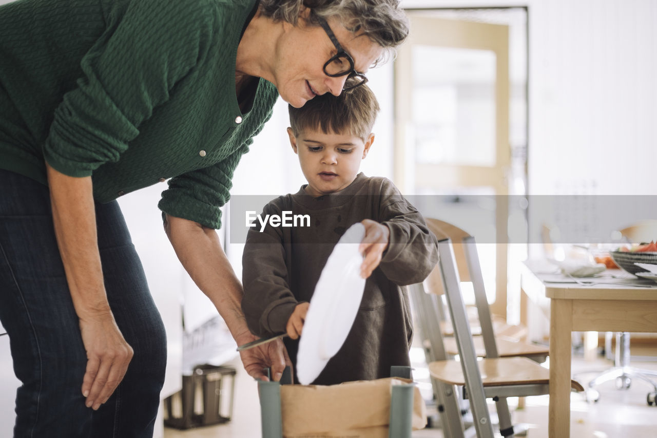 Boy throwing leftover food in garbage can with teacher at kindergarten