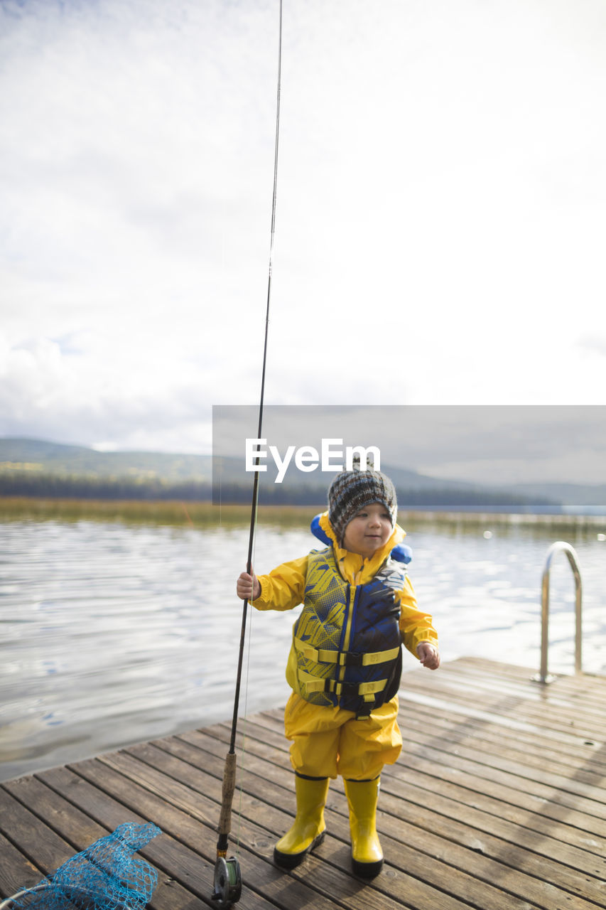 Baby boy wearing raincoat and life jacket while holding fishing rod on wooden pier over lake