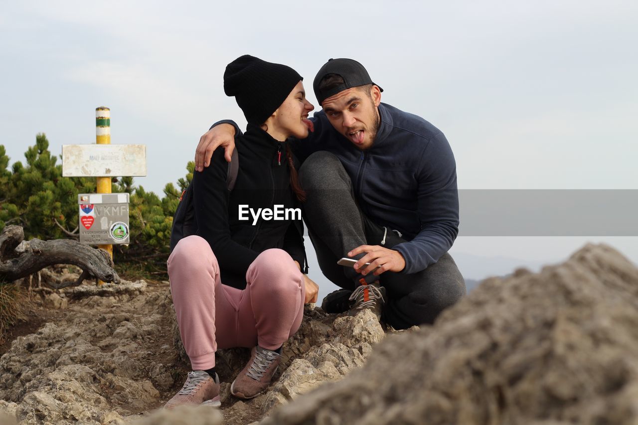 Friends sticking out tongues while sitting on rock at beach against sky