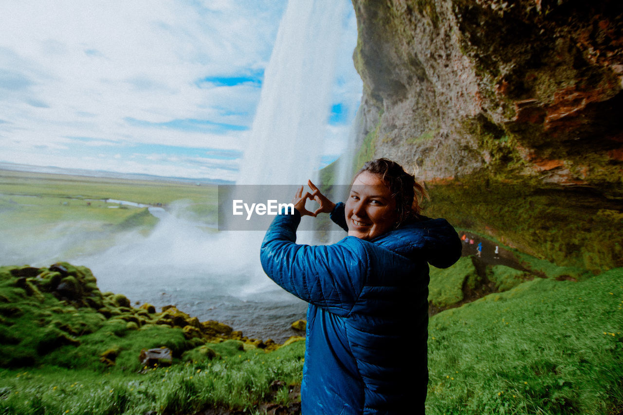 Portrait of smiling woman making heart shape against waterfall