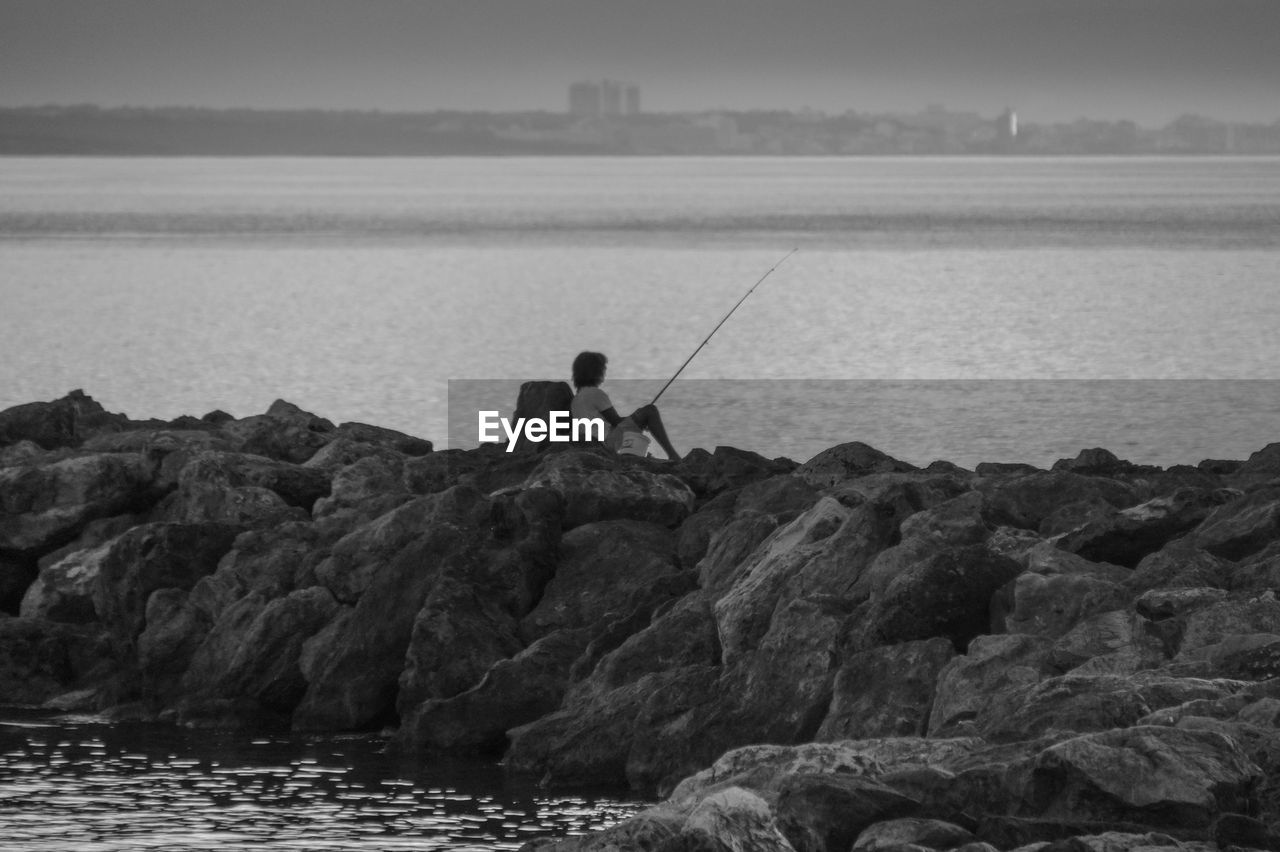Rear view of man fishing while sitting on rocks against sea