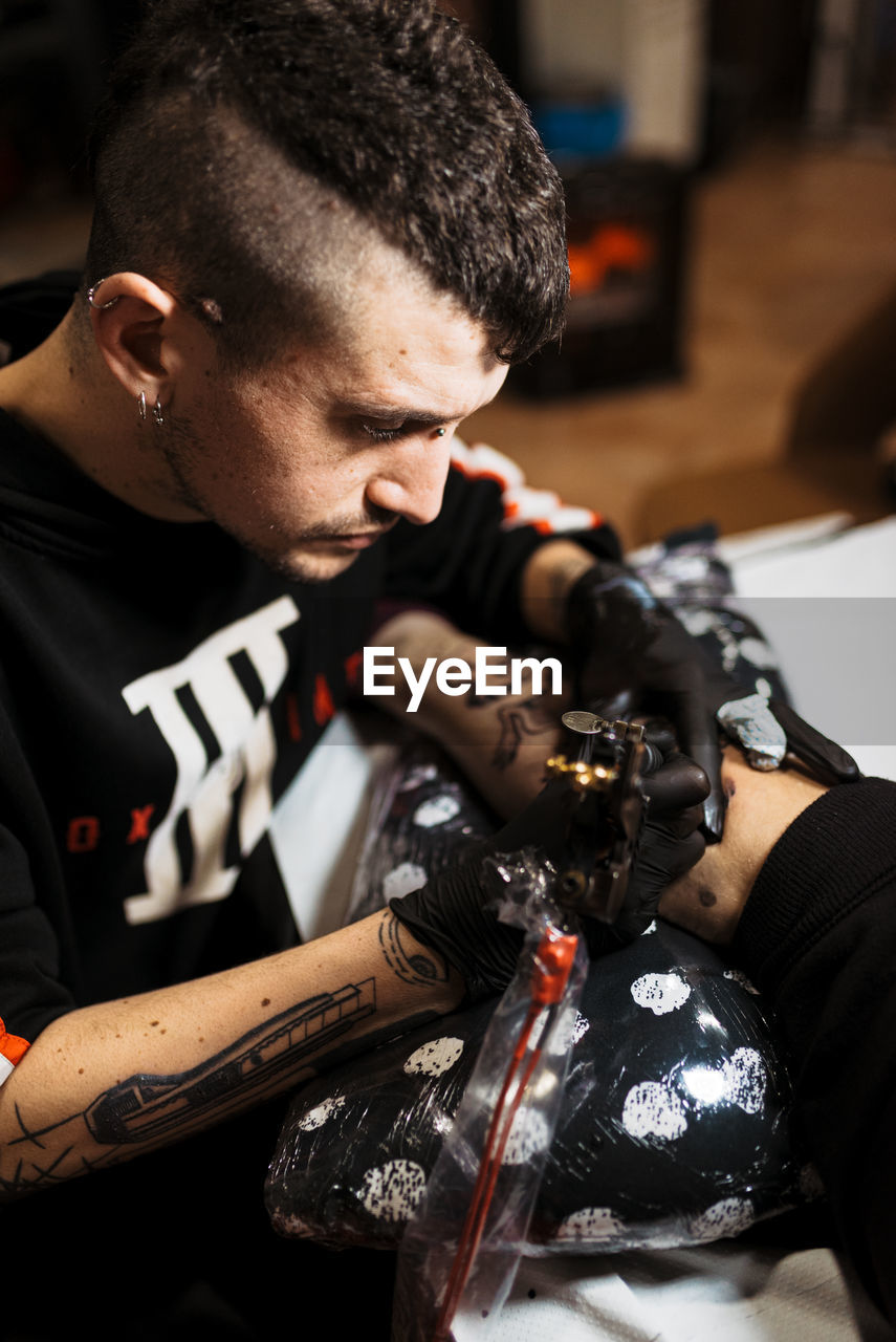 Stylish man with piercing using tattoo machine to make tattoo on leg of crop customer during work in salon