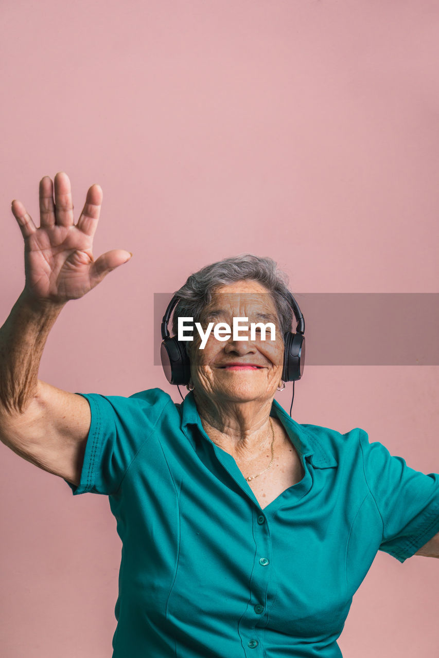 Joyful modern senior female listening to music in headphones and dancing with closed eyes on pink background in studio