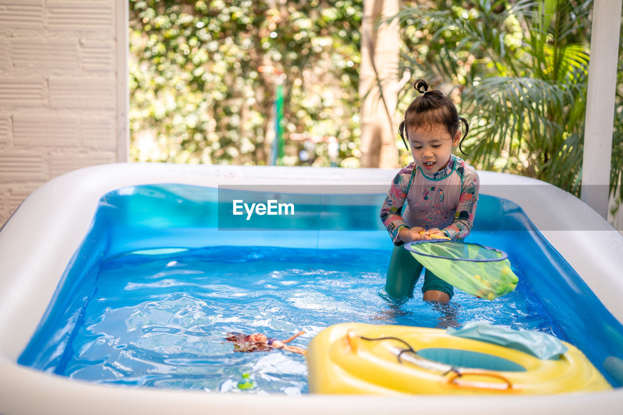 Cute little girl playing in an inflatable rubber swimming pool at home during quarantine period.