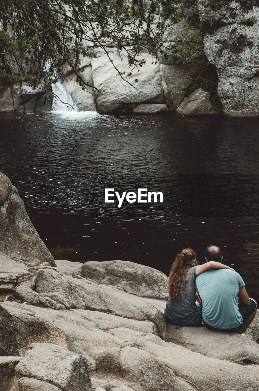 Rear view of friends sitting on rock while looking at lake