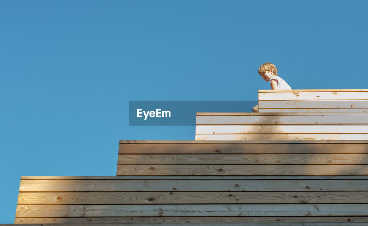 Low angle view of boy sitting on wooden structure against clear blue sky