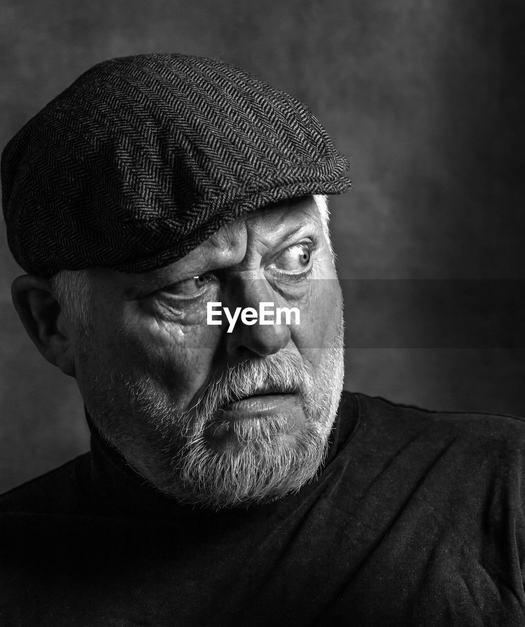 An elderly man with a beard and a cap looks to the side in shock. the picture is a  portrait.