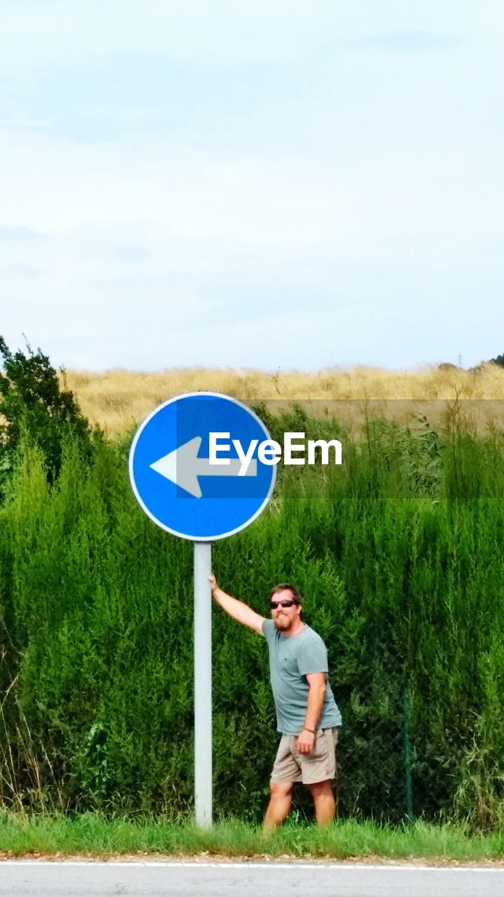 Man holding directional sign while standing on roadside against sky