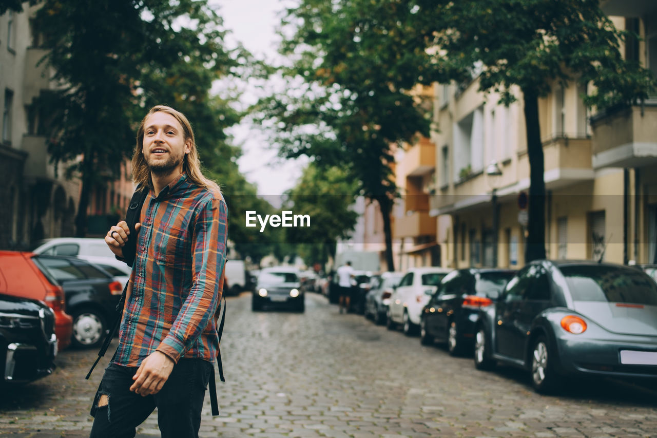 Young man carrying backpack while walking on cobbled street amidst cars in city