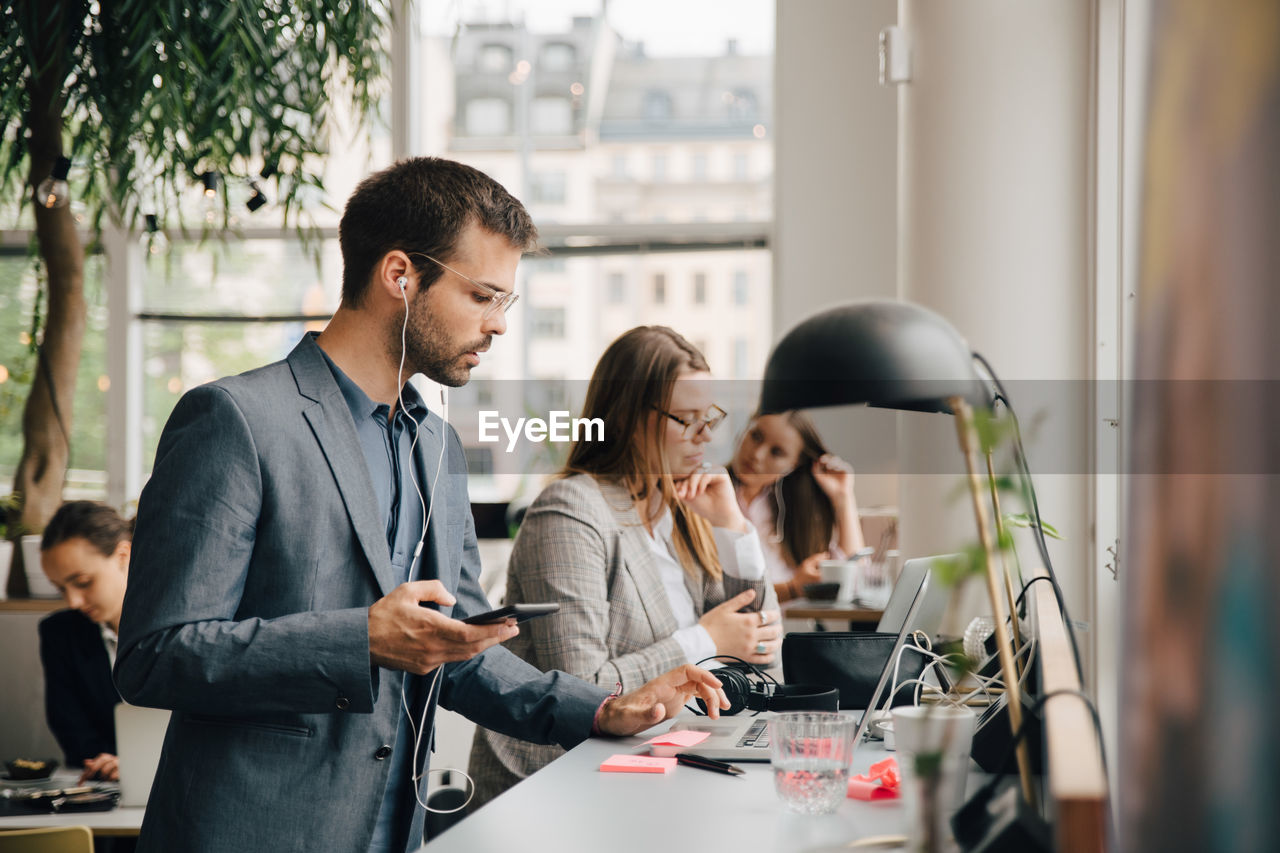 Male professional using laptop and smart phone while working with colleagues at coworking space