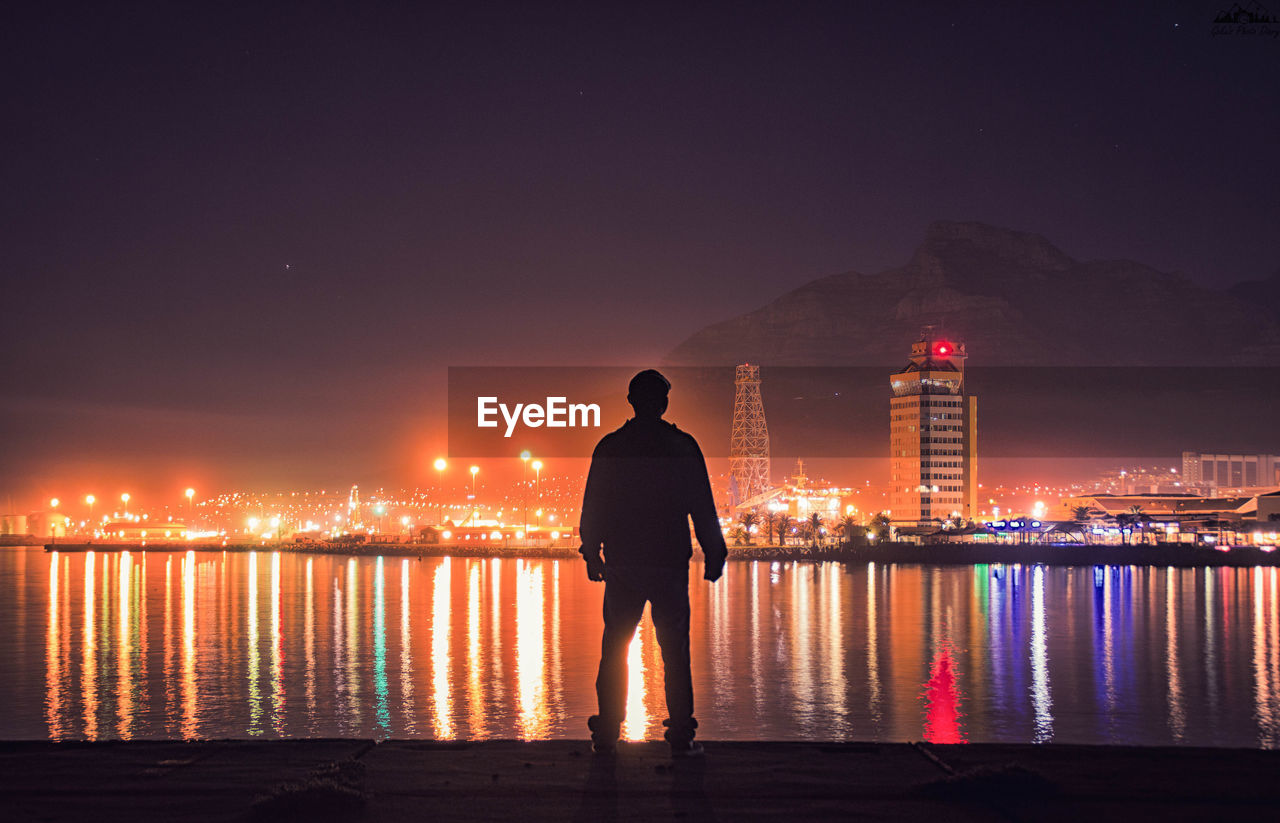 Silhouette man looking at view while standing by sea in illuminated city at night