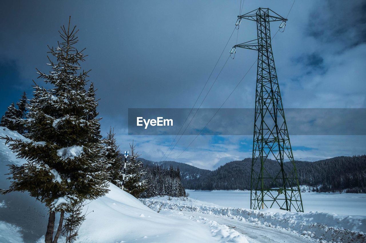 Electricity pylon on snow covered land against cloudy sky