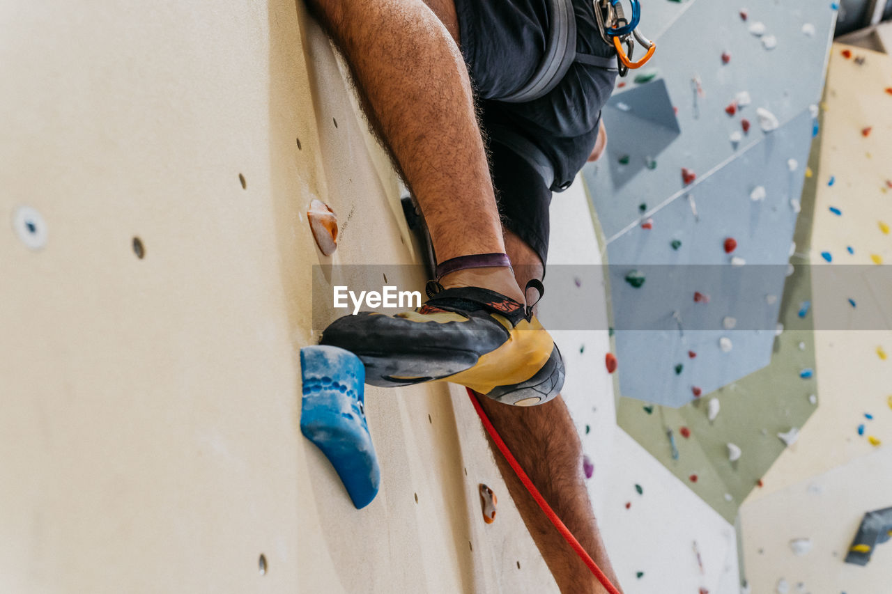 Crop anonymous sportive male alpinist with belay climbing up artificial wall with colorful grips during intense training in light gym