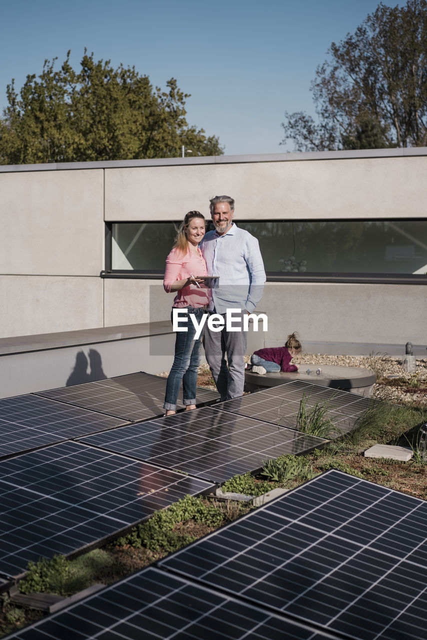 Smiling woman holding tablet pc standing with man amidst solar panel on sunny day