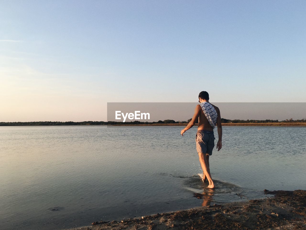 Rear view of shirtless man walking at lakeshore during sunset