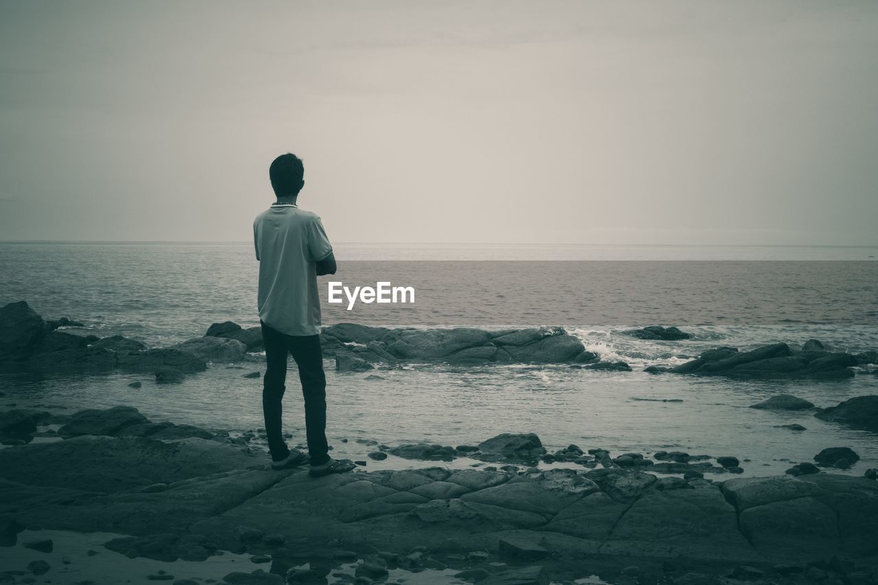 REAR VIEW OF MAN LOOKING AT SEA SHORE AGAINST SKY