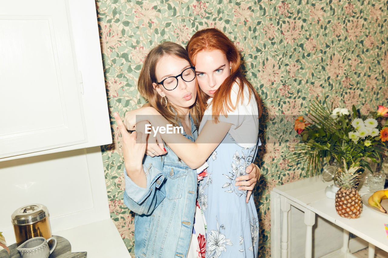 Portrait of young redhead woman embracing female friend showing horn sign at home