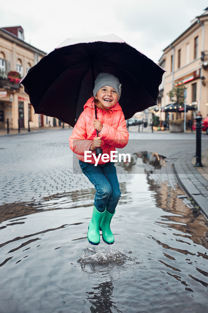 Happy smiling girl holding big umbrella jumping in the puddle during walk in a downtown