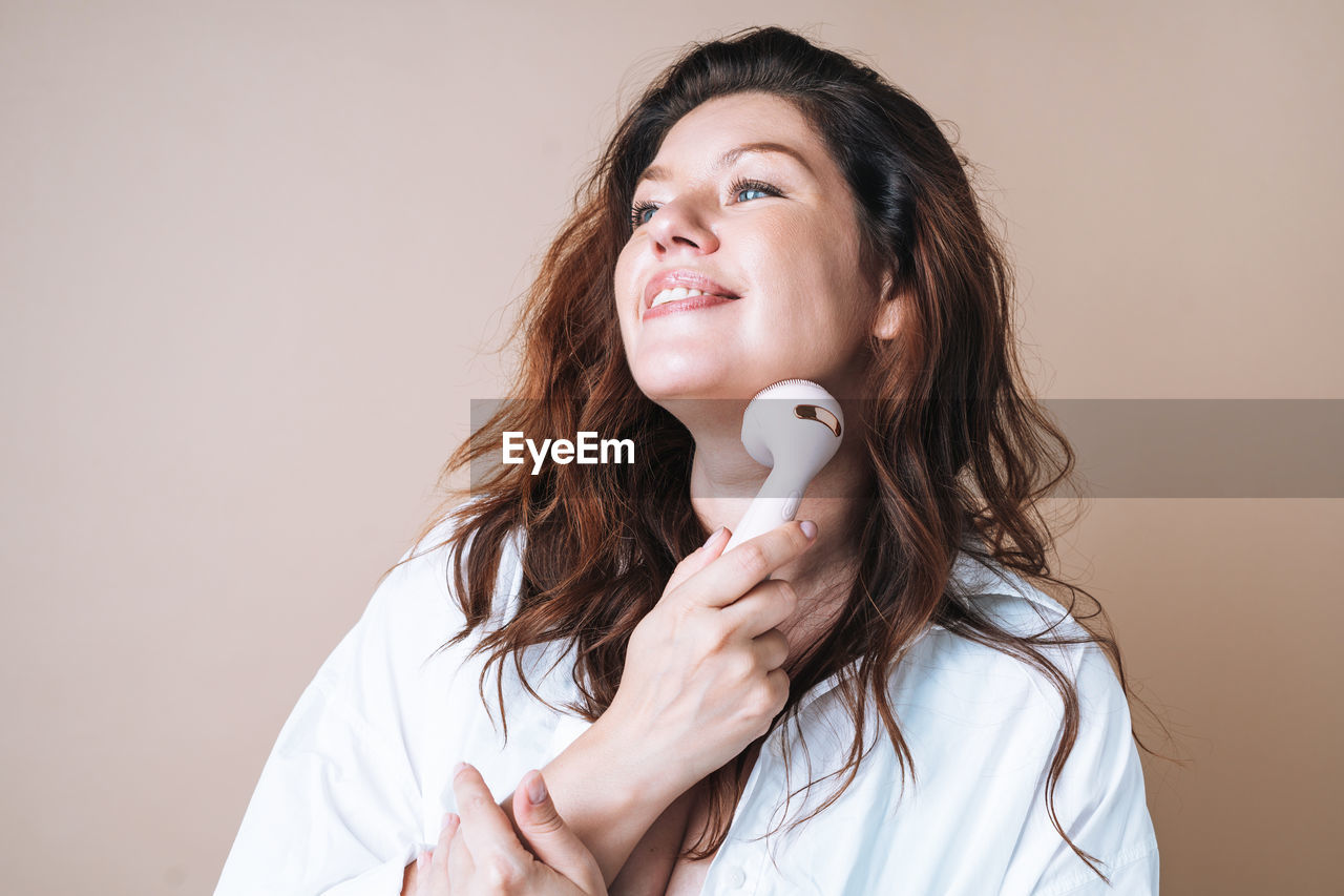 Self loving woman in white shirt with facial massager in hands on beige background, body love