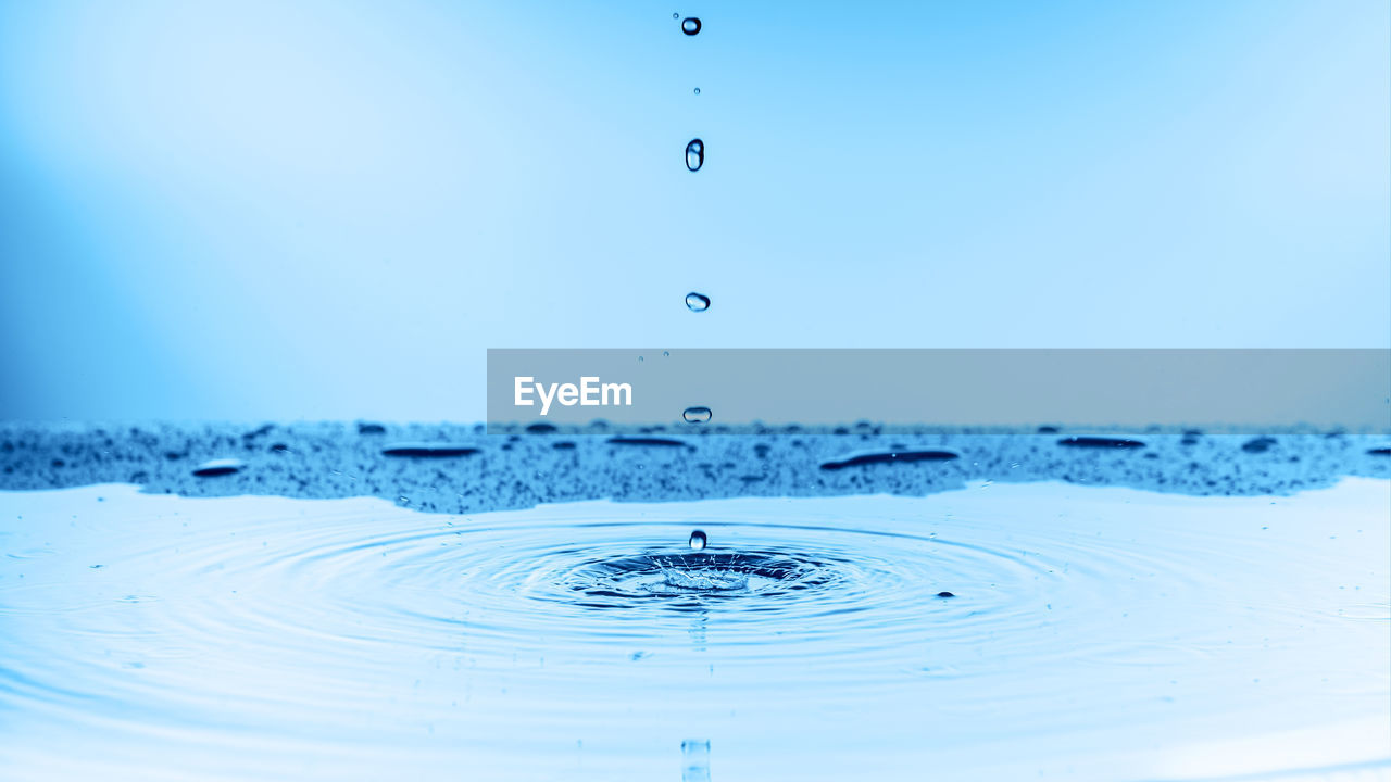 CLOSE-UP OF DROP FALLING ON BLUE WATER AGAINST WHITE BACKGROUND