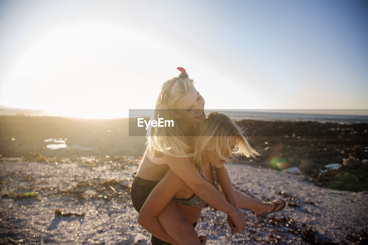 Young woman piggybacking cheerful female friend at beach during sunset